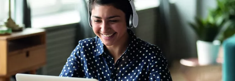 Smiling person wearing headphones and looking at a laptop in a cozy room.