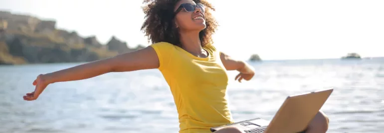 Woman in yellow shirt stretching on beach with laptop.