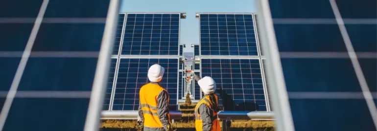 Two workers in hard hats and vests inspect large solar panels in a field.