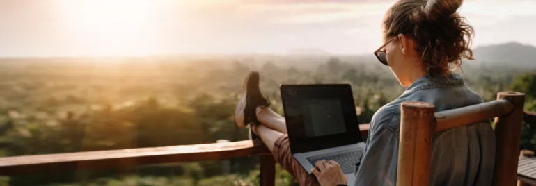 Person using a laptop on a balcony, overlooking a scenic sunset.