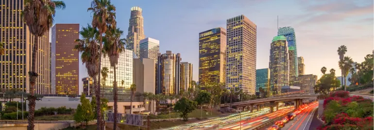 Los Angeles skyline at dusk with illuminated buildings and palm trees.