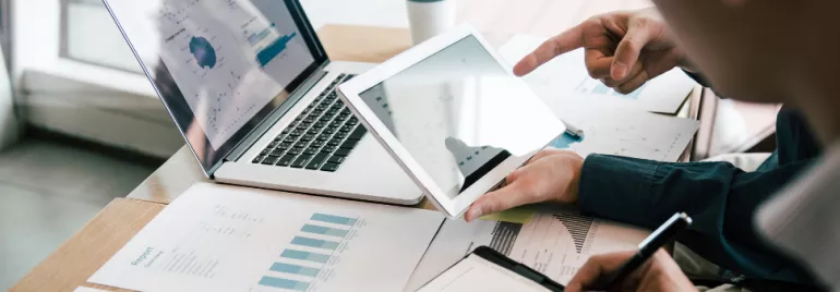 Two people reviewing financial graphs on a tablet and laptop at a desk.