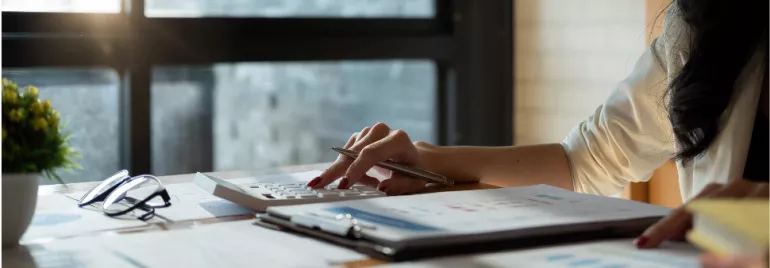 Person using a calculator at a desk with reports and glasses.