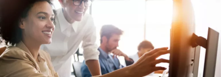 Two people smiling while working on a computer together in a bright office.