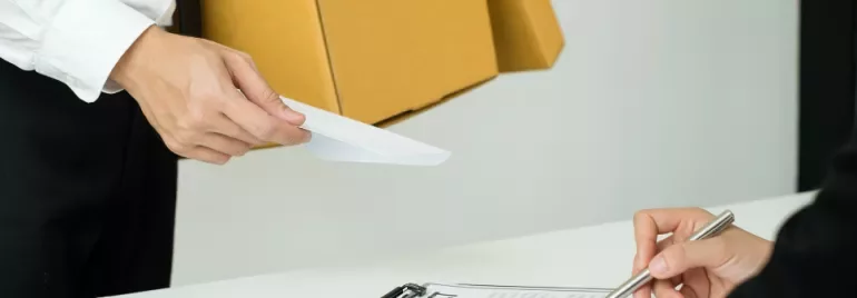 Person handing an envelope to someone signing a contract at a desk.