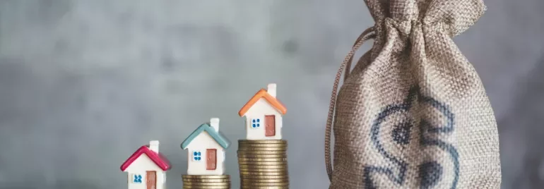 Three small house models on stacked coins beside a burlap sack with a dollar sign.