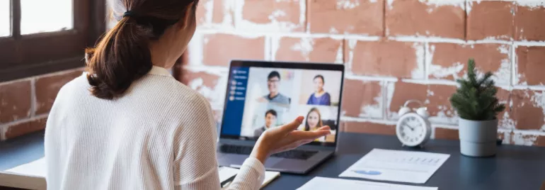 Person in a white sweater attending a video conference on a laptop at a desk with papers and a small plant.