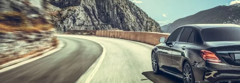 A sleek black car driving on a winding mountain road under a clear sky.