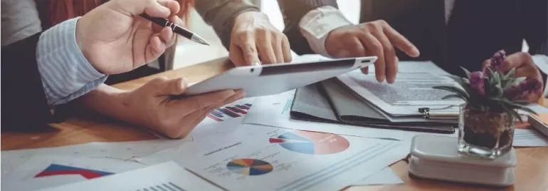 Hands holding a tablet over a table with charts and graphs.