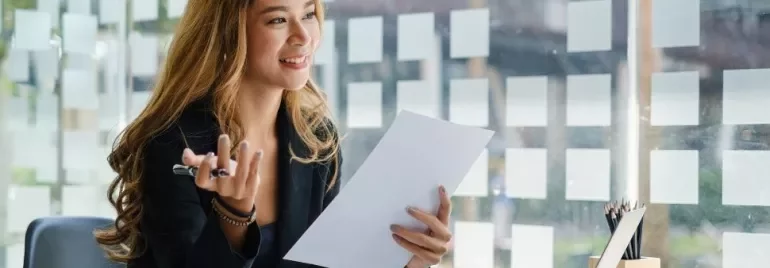 Smiling woman in an office holding a document, seated by a window with sticky notes.
