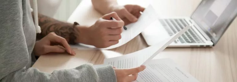 Two people reviewing documents with a laptop on a wooden desk