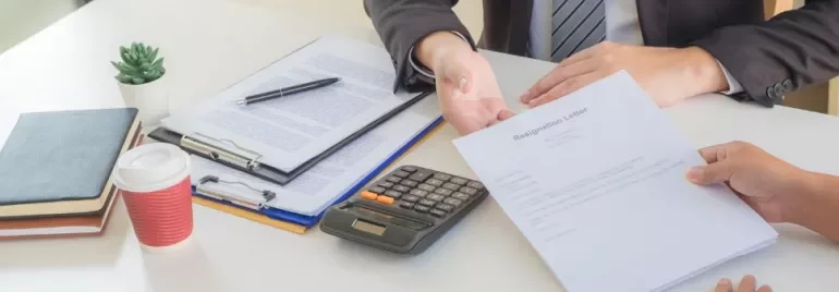 A person submits a resignation letter during a meeting with a colleague at a desk with a coffee and calculator.