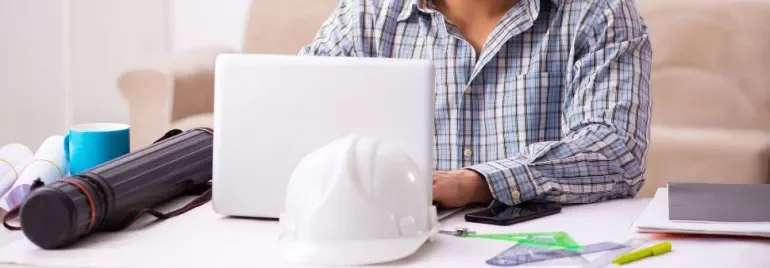 Man in plaid shirt working on a laptop with a white helmet on the table.