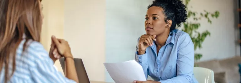 Two women having a focused discussion at a desk with a laptop and paperwork.