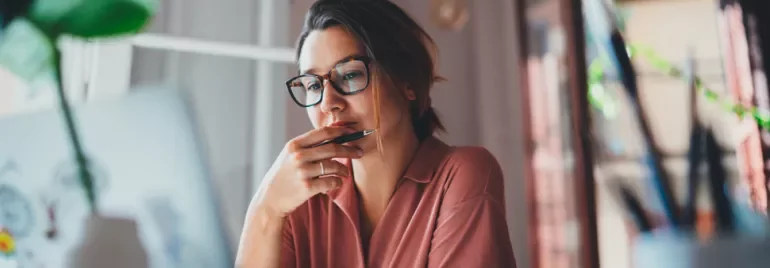 Woman in glasses working on a laptop, holding a pen to her chin and looking thoughtful.