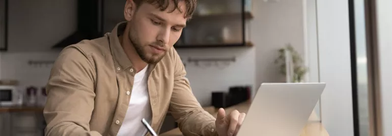 Man writing notes in front of a laptop at a home desk.