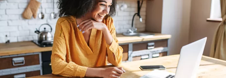 Smiling woman in yellow shirt using a laptop at a wooden table in a cozy kitchen.