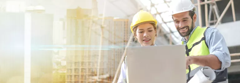 Two construction workers, a man and woman, reviewing plans on a laptop at a building site.