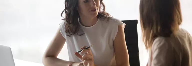 Two women having a conversation at a desk with a laptop nearby.