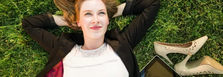 Woman lying on grass, smiling, with shoes and tablet nearby.