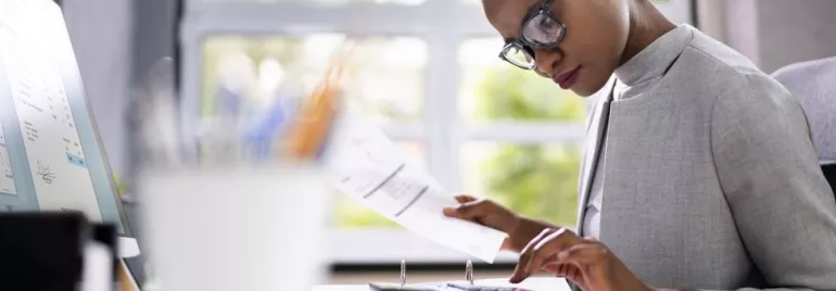 Woman calculating at desk with documents and monitor