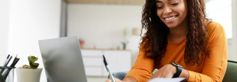 Woman in orange shirt writing on paper with laptop at table.