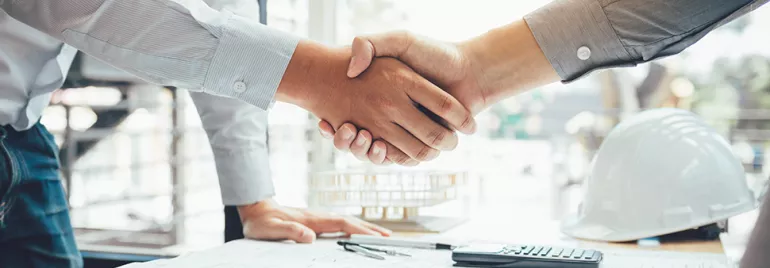Two people shaking hands over construction blueprints.