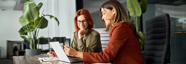 Two women discussing work at a desk in a modern office with large plants.
