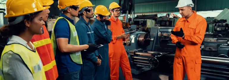 Factory workers in safety gear listen to a supervisor in an industrial setting.