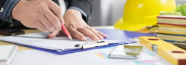 Person writing at a desk with construction tools and a yellow hard hat.