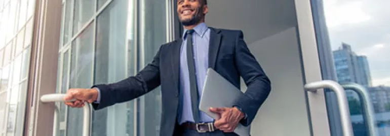 Businessman in a suit exiting a glass building, holding a laptop and smiling.
