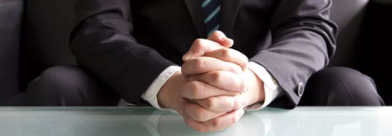 Person in suit sitting at a glass table with folded hands.