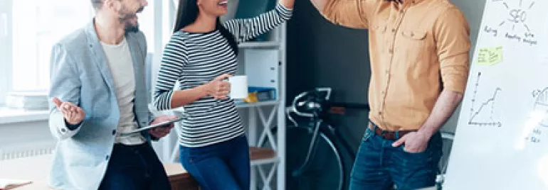 Three colleagues celebrate a success with a high-five next to a whiteboard in an office.