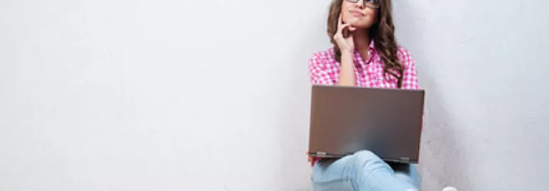 Woman sitting, thinking with a laptop on her lap against a white wall.