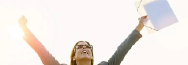 Person stands joyfully with arms raised, holding a folder against a bright sky.