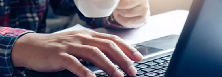 Person using a laptop while holding a coffee cup, sitting at a desk.