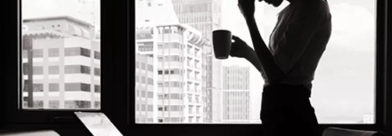 Silhouette of a woman holding a mug, standing by a window, overlooking city buildings, with laptops in the foreground.