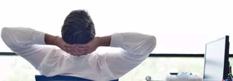 Person relaxed in office chair, hands behind head, near window.
