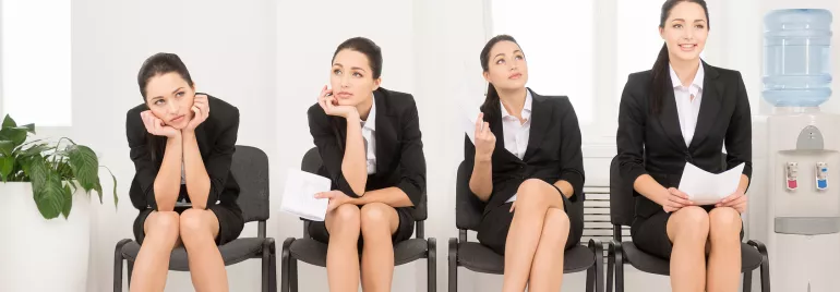 Four women in business attire sitting on chairs with different expressions, next to a water cooler.