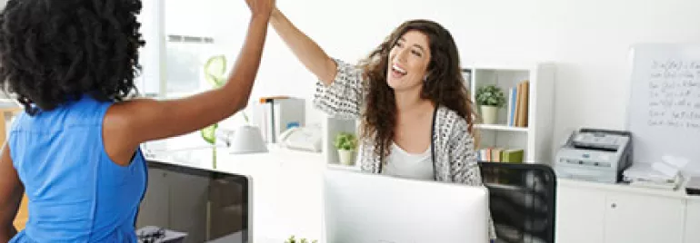 Two women in an office giving a high-five, smiling at a desk with computers.