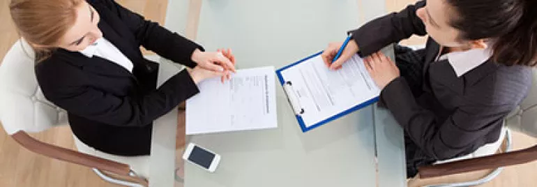 Two businesswomen at a table, one taking notes on a clipboard.