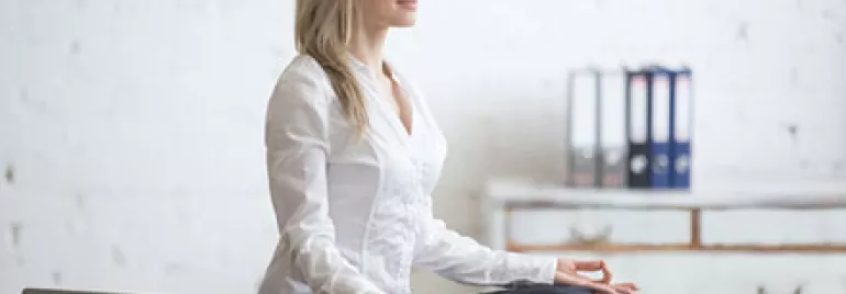 Woman in business attire meditating cross-legged on a desk in a bright office.
