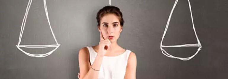 Woman pondering, standing in front of sketched balance scales on a blackboard.