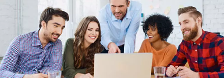 Group of colleagues smiling and discussing around a laptop in a bright office.