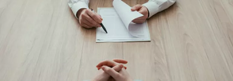 Two people sitting at a table, one holding a pen and paper, in an office setting.