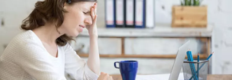Woman holding head in frustration at desk with laptop and coffee.