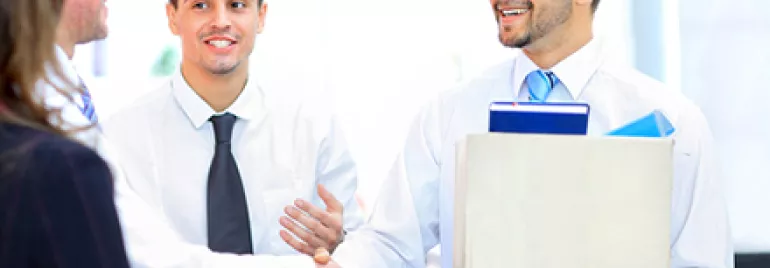 A man holding a box is smiling and shaking hands with another man in an office setting.