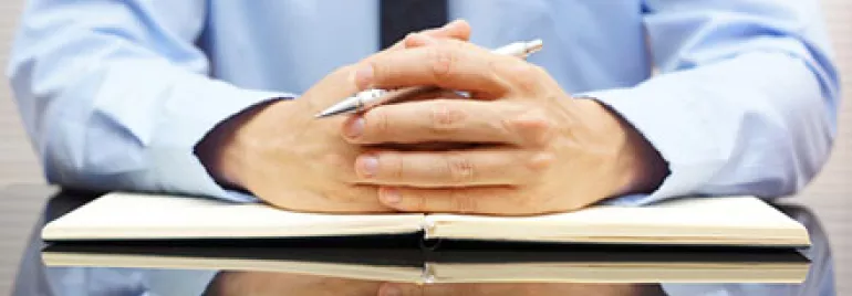Person in a blue shirt sitting at a desk with a notebook and pen.