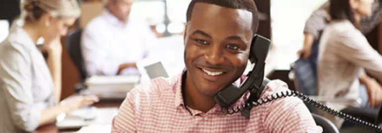 Man smiling while talking on the phone in a busy office.