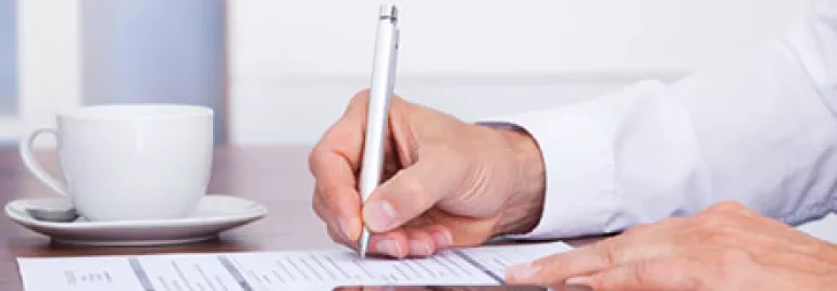 Person signing a paper at a desk with a pen and smartphone nearby.